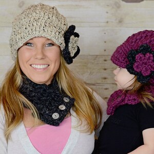 May include: Two crocheted hats with matching scarves. The hat on the left is beige with a black flower, and the hat on the right is burgundy with a black flower. Both hats have a slouchy style.