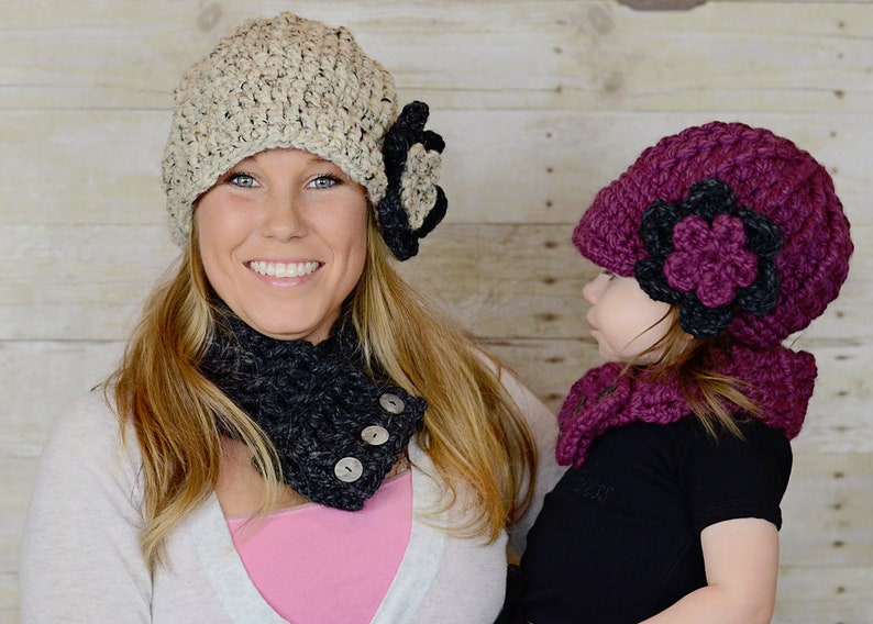 May include: Two crocheted hats with matching scarves. The hat on the left is beige with a black flower, and the hat on the right is burgundy with a black flower. Both hats have a slouchy style.