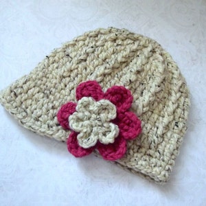 Puede incluir: Un gorro de punto de color crema con un adorno de flor rosa y blanca.