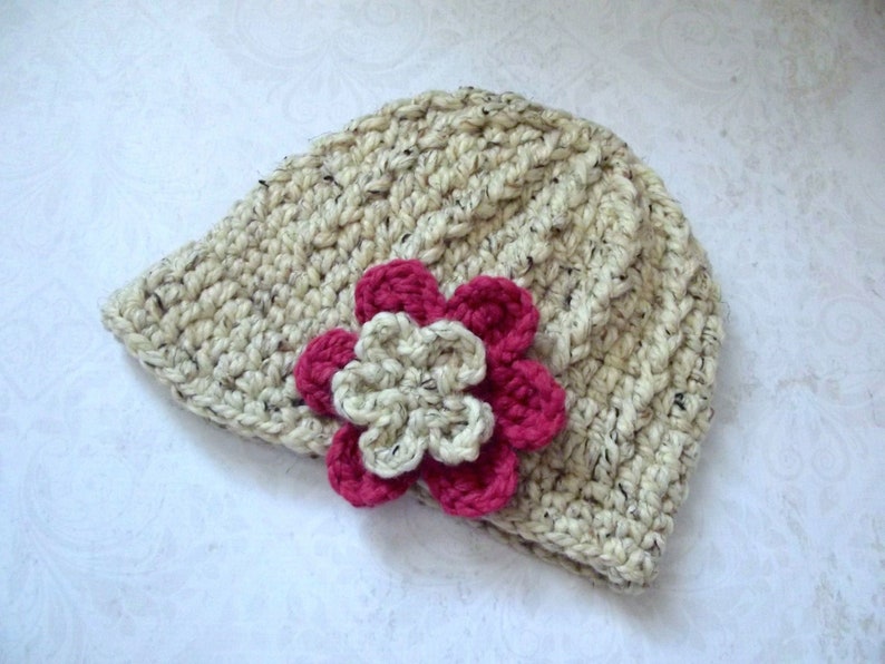 Puede incluir: Un gorro de punto de color crema con un adorno de flor rosa y blanca.