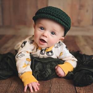 May include: A baby wearing a green knitted cap and a white onesie with a black and white animal print. The baby is lying on a green fabric.