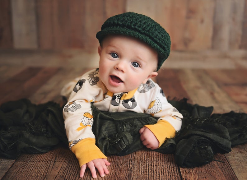May include: A baby wearing a green knitted cap and a white onesie with a black and white animal print. The baby is lying on a green fabric.