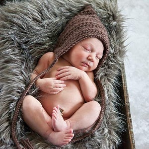 May include: A newborn baby wearing a brown crocheted pixie hat, lying peacefully on a faux fur blanket inside a wooden box. The baby is curled up with hands crossed over the chest.