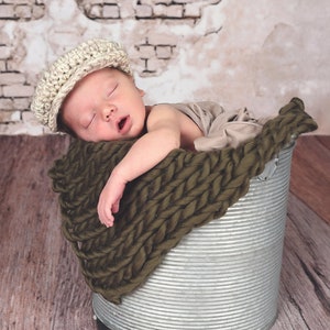 May include: A newborn baby wearing a beige knitted cap sleeps in a metal bucket lined with a dark green knitted blanket.