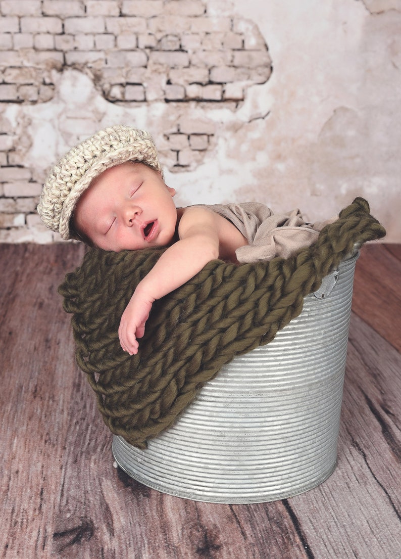 May include: A newborn baby wearing a beige knitted cap sleeps in a metal bucket lined with a dark green knitted blanket.