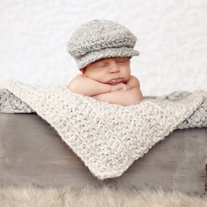 May include: A newborn baby wearing a gray knitted cap sleeps in a wooden crate lined with a white crocheted blanket.