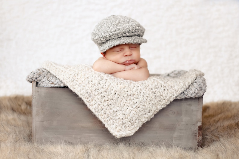 May include: A newborn baby wearing a gray knitted cap sleeps in a wooden crate lined with a white crocheted blanket.