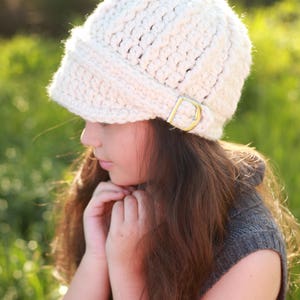 May include: A white crocheted hat with a wide brim and a buckle detail. The hat is being worn by a person with long brown hair.