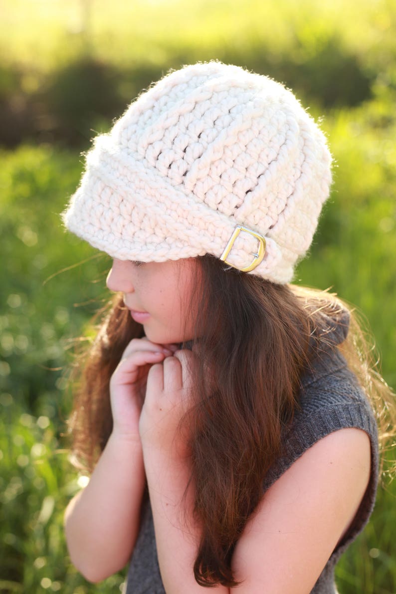 May include: A white crocheted hat with a wide brim and a buckle detail. The hat is being worn by a person with long brown hair.