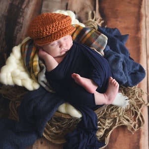 May include: A newborn baby wearing a brown crocheted cap and a navy blue swaddle is sleeping in a woven basket with a white, cream, and plaid blanket.