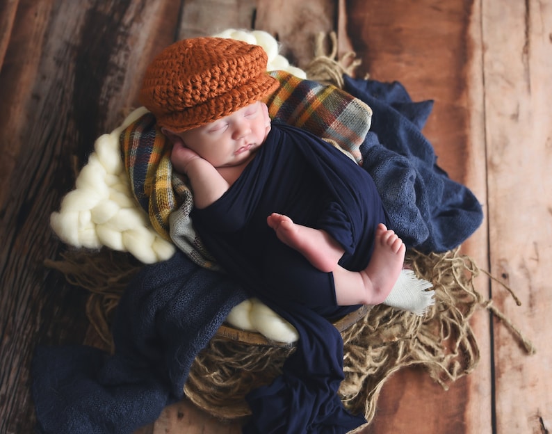 May include: A newborn baby wearing a brown crocheted cap and a navy blue swaddle is sleeping in a woven basket with a white, cream, and plaid blanket.