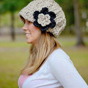May include: A woman wearing a beige crocheted beanie with a black and beige crocheted flower accent. She is wearing a white cardigan and has long blonde hair.