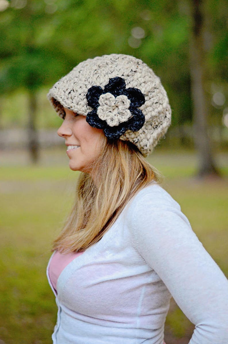 May include: A woman wearing a beige crocheted beanie with a black and beige crocheted flower accent. She is wearing a white cardigan and has long blonde hair.