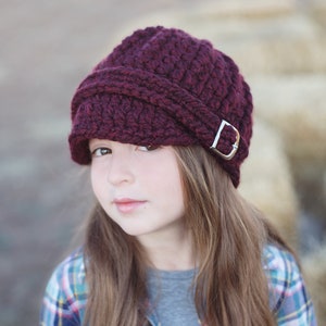 May include: A burgundy crocheted newsboy hat with a silver buckle. The hat is worn by a person with long brown hair.