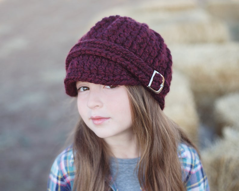May include: A burgundy crocheted newsboy hat with a silver buckle. The hat is worn by a person with long brown hair.