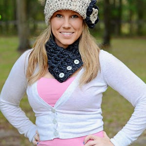 May include: A woman wearing a white cardigan, a pink shirt, blue jeans, a crocheted hat with a flower accent, and a black crocheted neck warmer with buttons.