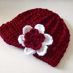 Puede incluir: Un gorro de punto de color burdeos con una flor de ganchillo blanca en el lateral.