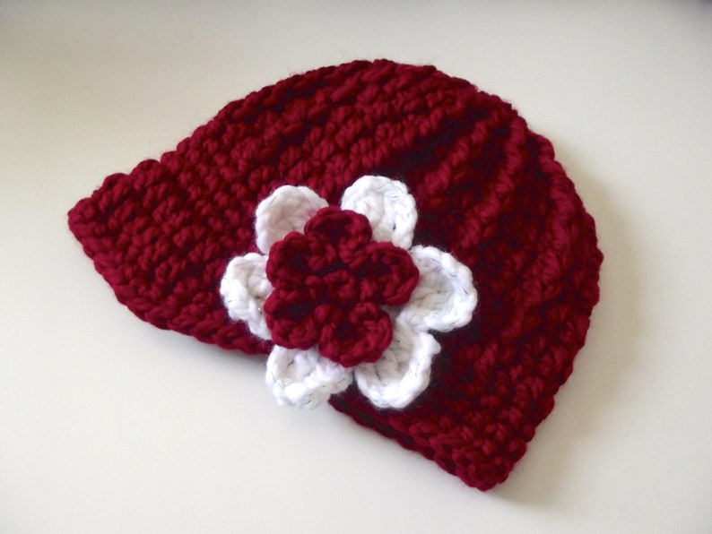 Puede incluir: Un gorro de punto de color burdeos con una flor de ganchillo blanca en el lateral.
