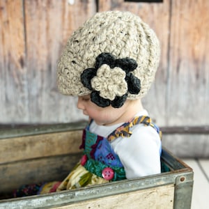 May include: A young child wearing a beige crocheted hat with a black flower accent sits in a wooden crate. The hat has a unique textured design and the flower is a statement piece.