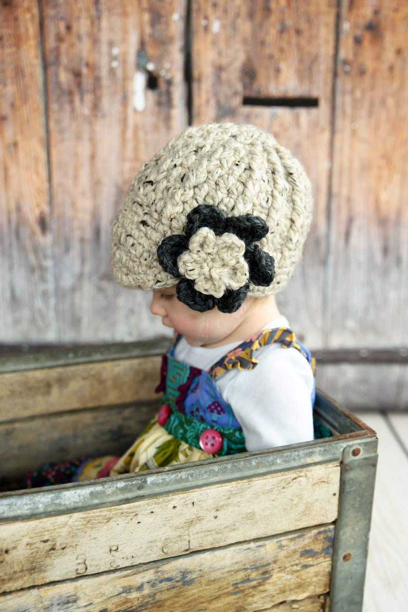 May include: A young child wearing a beige crocheted hat with a black flower accent sits in a wooden crate. The hat has a unique textured design and the flower is a statement piece.