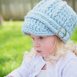 May include: A young child wearing a light blue crocheted hat with a silver buckle. The hat has a slouchy, relaxed fit and covers the child's forehead. The child is wearing a white shirt and has long blonde hair.