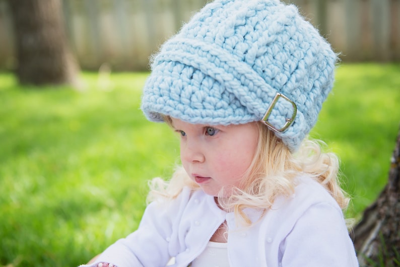 May include: A young child wearing a light blue crocheted hat with a silver buckle. The hat has a slouchy, relaxed fit and covers the child's forehead. The child is wearing a white shirt and has long blonde hair.