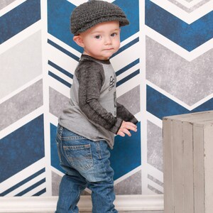 May include: A young child wearing a gray newsboy cap, a gray and black long-sleeved shirt, blue jeans, and black sneakers. The child is standing in front of a blue and gray chevron patterned wall.