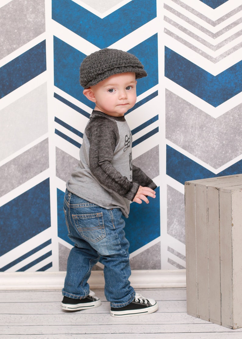 May include: A young child wearing a gray newsboy cap, a gray and black long-sleeved shirt, blue jeans, and black sneakers. The child is standing in front of a blue and gray chevron patterned wall.