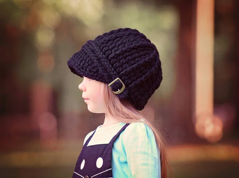 May include: A black knitted beanie with a fold-down brim and a metal buckle. The beanie is worn by a young person with long brown hair.