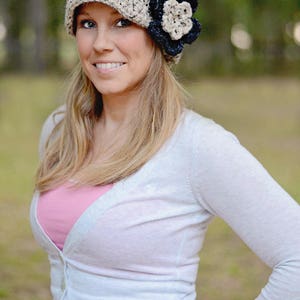 May include: A woman wearing a beige crocheted beanie with a black and white flower accent. She is wearing a white cardigan over a pink shirt.
