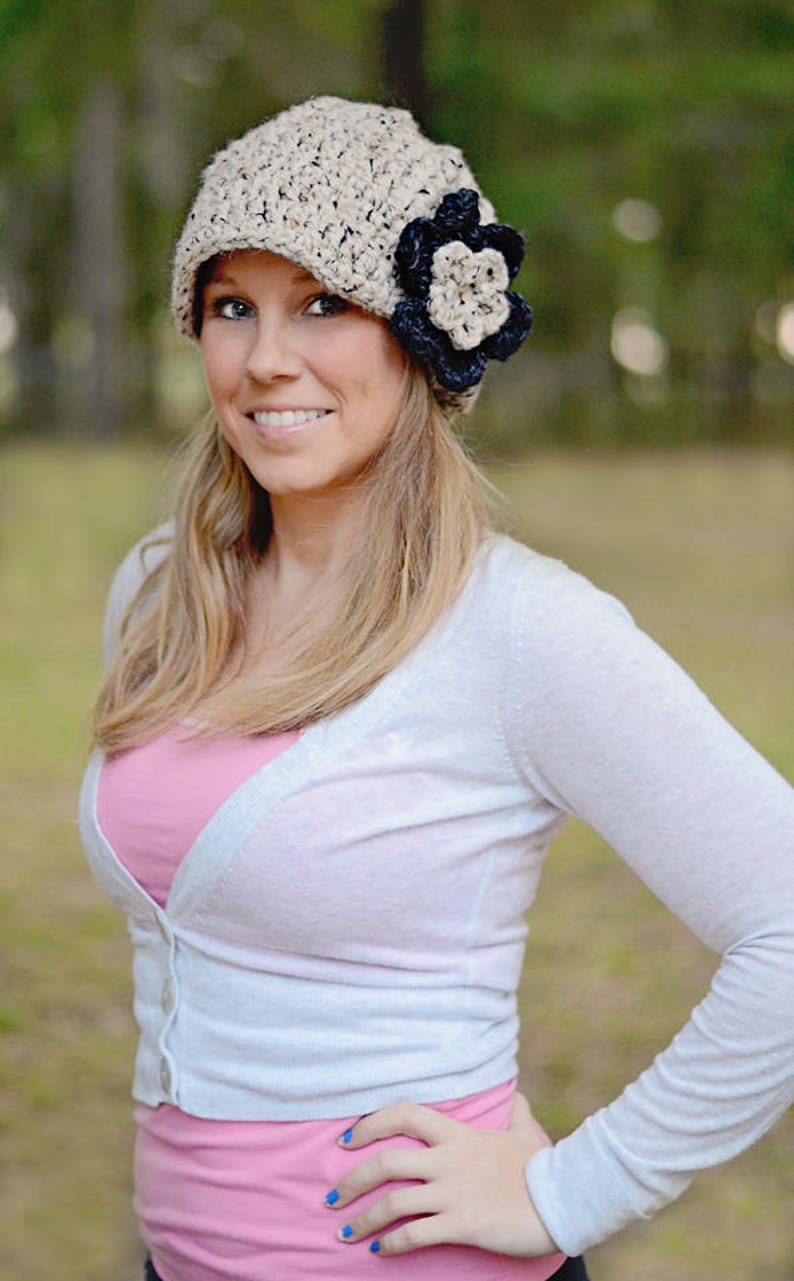 May include: A woman wearing a beige crocheted beanie with a black and white flower accent. She is wearing a white cardigan over a pink shirt.