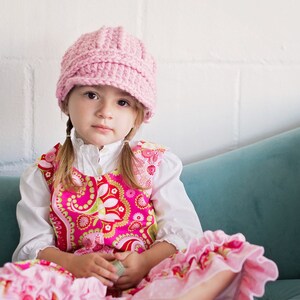 May include: A young girl wearing a pink crocheted hat and a pink and red floral dress sits on a teal sofa.