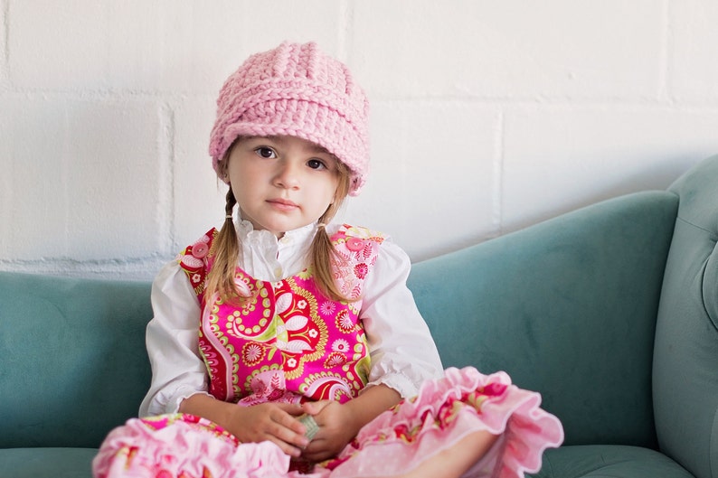 May include: A young girl wearing a pink crocheted hat and a pink and red floral dress sits on a teal sofa.