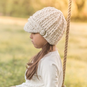 May include: A young person wearing a white crocheted beanie with a brim and a metal buckle. The beanie is worn on their head and they are looking down.