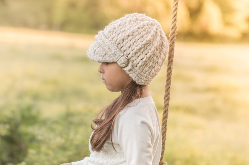 May include: A young person wearing a white crocheted beanie with a brim and a metal buckle. The beanie is worn on their head and they are looking down.