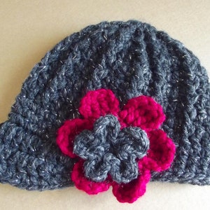 Puede incluir: Un gorro de punto gris con un acabado brillante y una gran flor rosa como adorno.