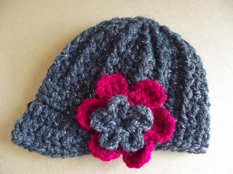 Puede incluir: Un gorro de punto gris con un acabado brillante y una gran flor rosa como adorno.