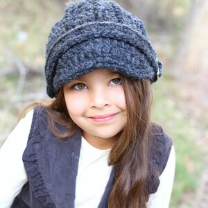 May include: A young person wearing a grey crocheted newsboy cap with a black waistcoat and a white shirt. The person has long brown hair and is smiling.