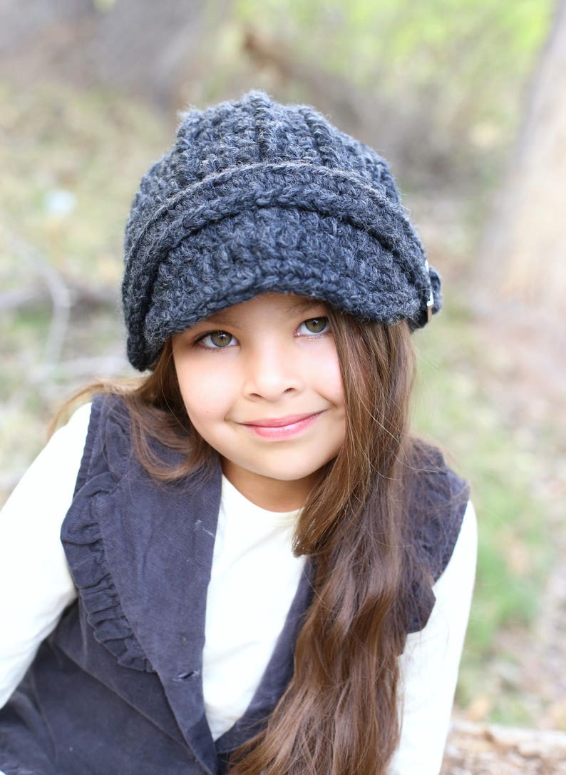 May include: A young person wearing a grey crocheted newsboy cap with a black waistcoat and a white shirt. The person has long brown hair and is smiling.