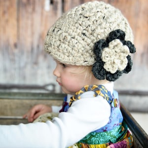 May include: A young child wearing a beige crocheted hat with a black crocheted flower accent. The hat is worn on the child's head, and the child is looking to the side. The child is wearing a colorful patterned dress.