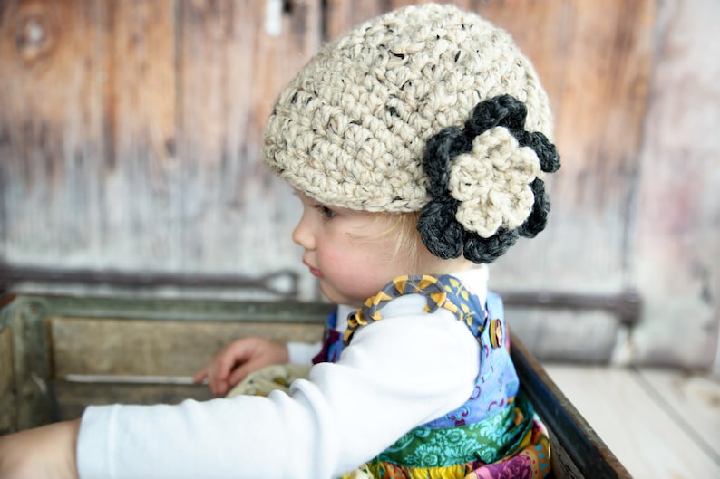May include: A young child wearing a beige crocheted hat with a black crocheted flower accent. The hat is worn on the child's head, and the child is looking to the side. The child is wearing a colorful patterned dress.