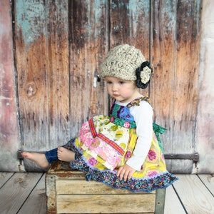 May include: A young child wearing a beige crocheted hat with a black flower accent sits on a wooden crate. The child is wearing a colorful patchwork dress with a white long-sleeved shirt. The child's feet are bare and the background is a weathered wooden door.