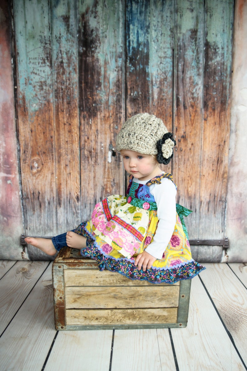 May include: A young child wearing a beige crocheted hat with a black flower accent sits on a wooden crate. The child is wearing a colorful patchwork dress with a white long-sleeved shirt. The child's feet are bare and the background is a weathered wooden door.