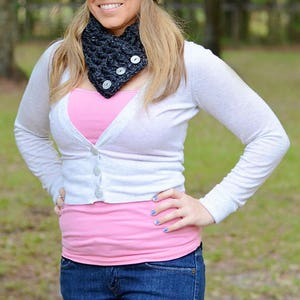 May include: A woman wearing a white cardigan, a pink tank top, blue jeans, a black and white crocheted hat with a flower, and a black crocheted neck warmer.