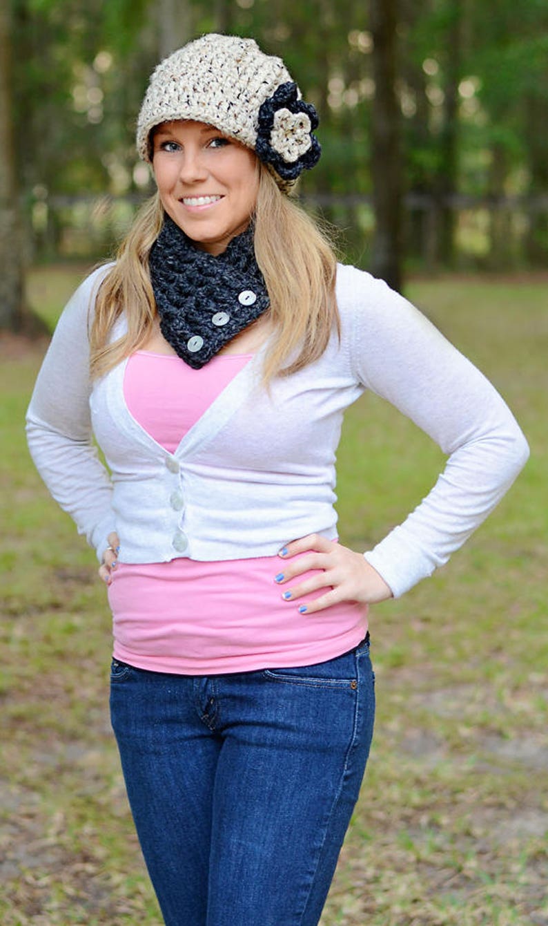 May include: A woman wearing a white cardigan, a pink tank top, blue jeans, a black and white crocheted hat with a flower, and a black crocheted neck warmer.