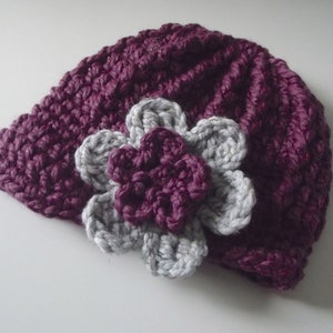 Puede incluir: Un gorro de punto morado con una flor de punto gris en la parte superior.