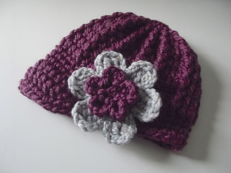 Puede incluir: Un gorro de punto morado con una flor de punto gris en la parte superior.