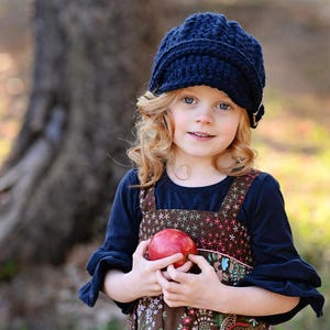 May include: A young girl with curly blonde hair wears a blue knitted hat and a brown dress with a floral pattern. She is holding a red apple in her hands.