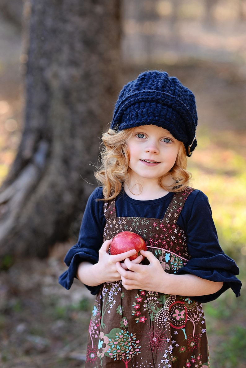 May include: A young girl with curly blonde hair wears a blue knitted hat and a brown dress with a floral pattern. She is holding a red apple in her hands.