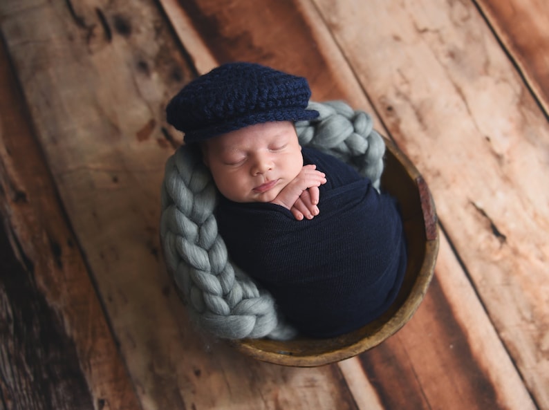 May include: A newborn baby wearing a blue knitted cap is sleeping in a woven basket. The baby is wrapped in a blue blanket and is lying on a light blue braided rope.